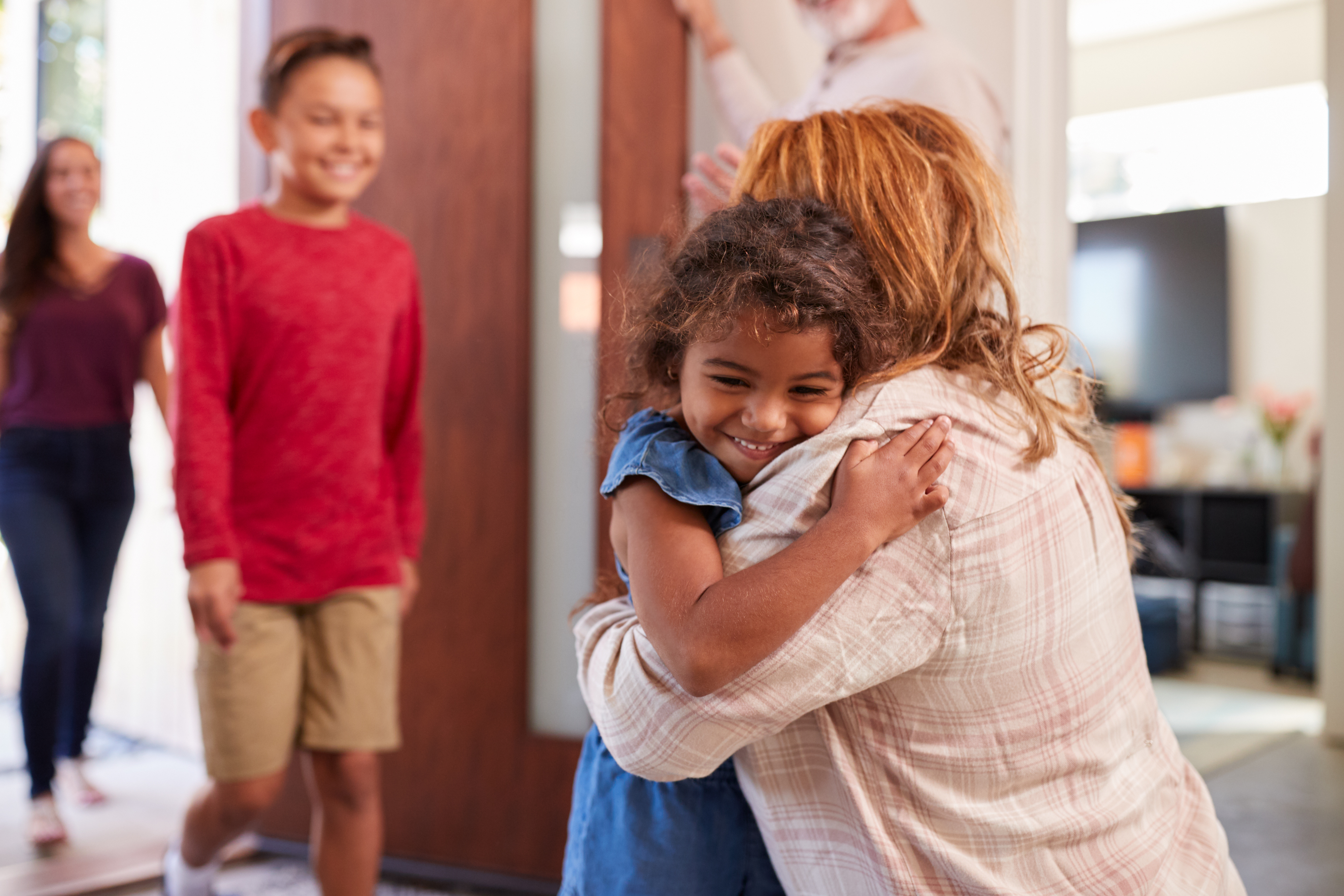an adult woman bent over hugging an female child, an adolescent boy and adult woman behind them walking through a doorway
