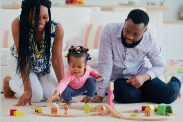 Adult woman and man sitting no floor with little girl between them playing with wooden toys.