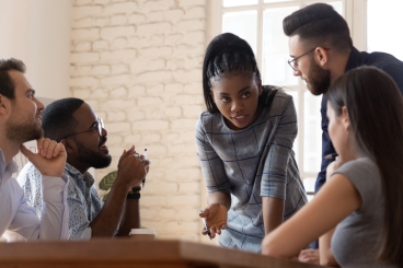 Female employee leaning over conference table while speaking to four coworkers.