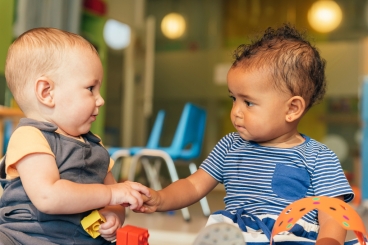 Two babies looking and each other and holding hands while playing with blocks.