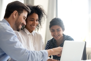 Two women and a male look at the laptop screen while the man points to the screen.