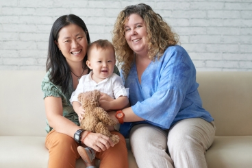 Two women holding baby while sitting on sofa, smiling.