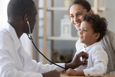 An adult male putting a stethoscope on a young child who is sitting in the lap of an adult woman.