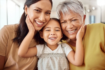 a female child sitting between an adult female and a female grandparent with her hands raised up and placed on the cheeks